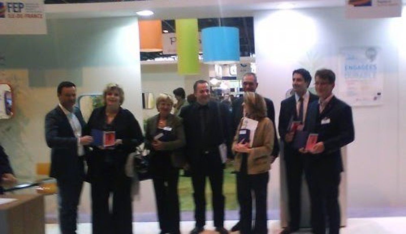 Huit personnes posent ensemble sur un stand de salon en tenant des livrets rouges, sous des lampes suspendues colorées.