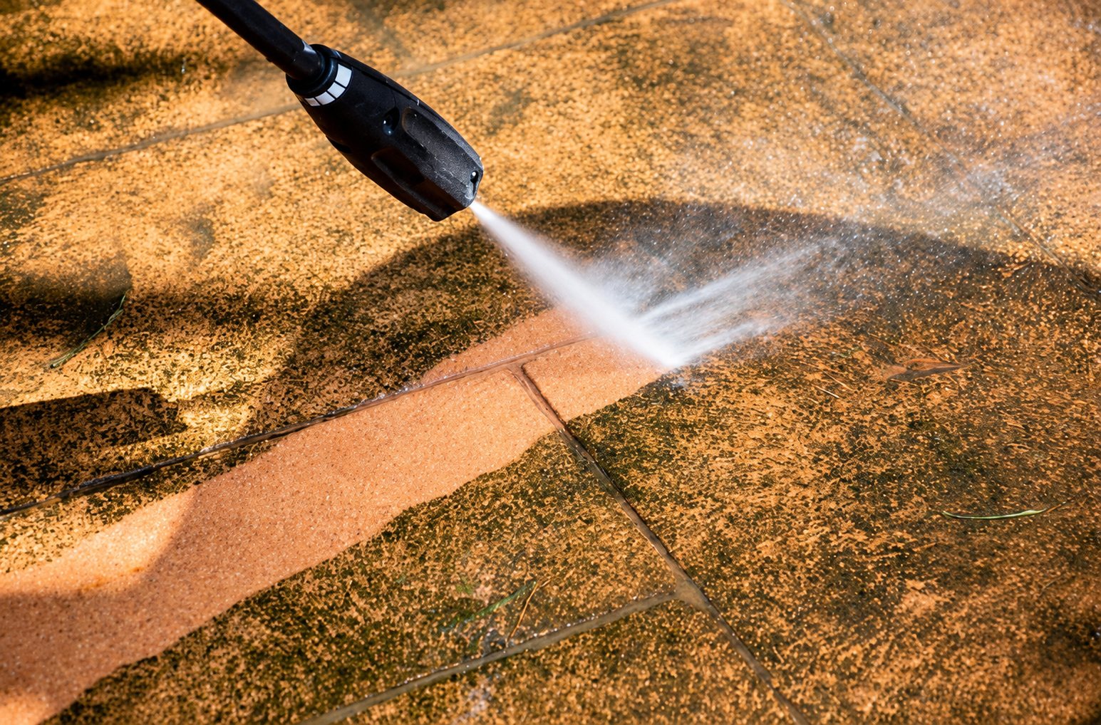 Une lance de nettoyeur haute pression projette un jet d'eau sur des dalles de terrasse, laissant apparaître une bande nettoyée plus claire au milieu des zones sales.